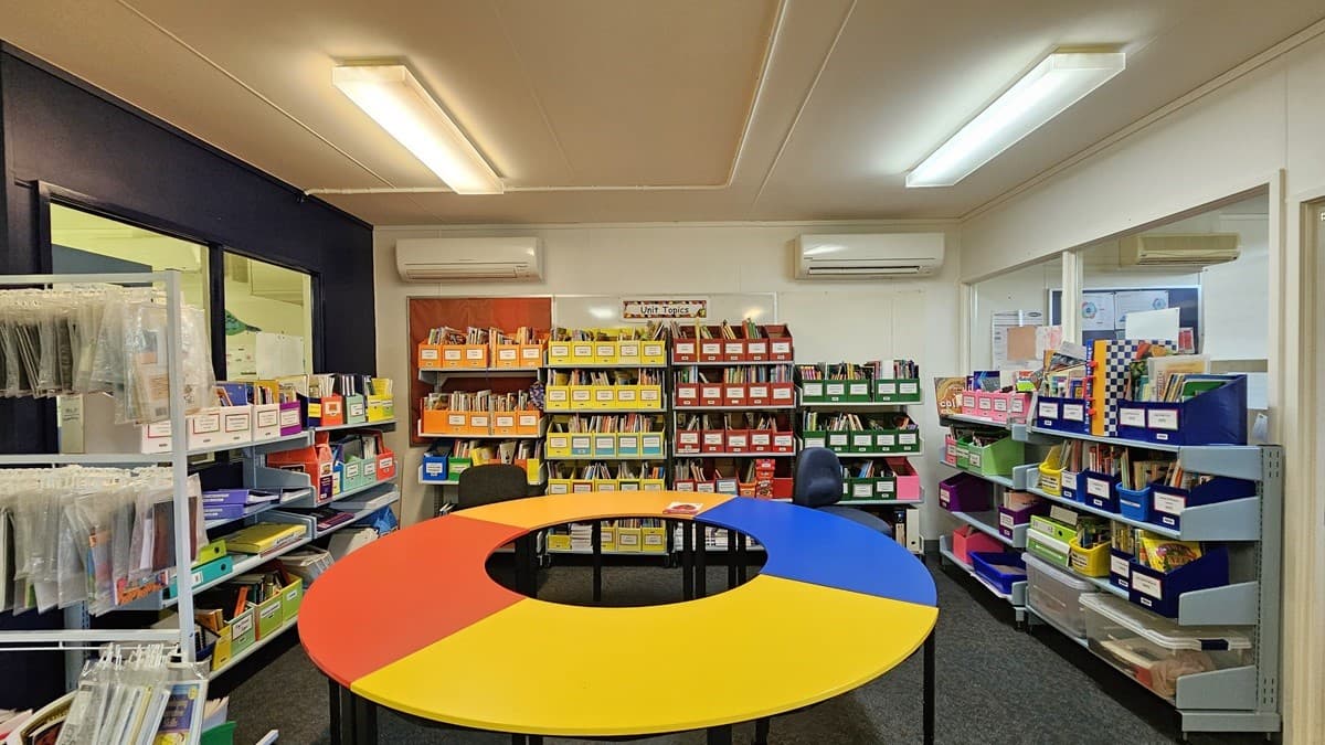 On the left, there are shelves filled with various educational materials and a dark blue wall with a window. In the middle, there is a round table with red, yellow, and blue sections, surrounded by chairs. On the right, more shelves hold colourful boxes and books. The background features additional shelves with neatly organised books and materials. The floor is carpeted in grey, and the ceiling is white with fluorescent light fixtures.
