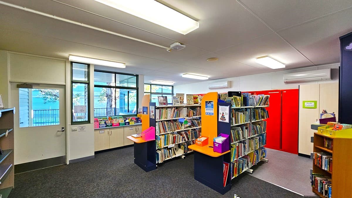 On the left, there are windows with a view of trees and a blue building, along with a white door. In the middle, there are bookshelves filled with books and colourful bins. On the right, there are more bookshelves, a bright red double door, and a small wooden shelf with additional books. The background includes more windows and a white wall. The floor is carpeted in grey, and the ceiling is white with fluorescent light fixtures.