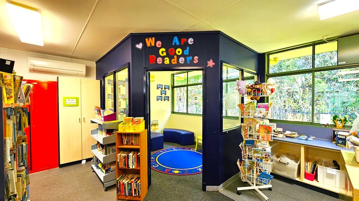 On the left, there are tall bookshelves filled with books and a red door next to a white cabinet. In the middle, there is a small reading nook with a blue circular rug and a few cushions, surrounded by dark blue walls with colourful letters spelling "We Are Good Readers." On the right, there are large windows with a view of trees, a white shelf with books and educational materials, and a rotating display rack. The floor is carpeted in grey, and the ceiling is white with fluorescent lights.