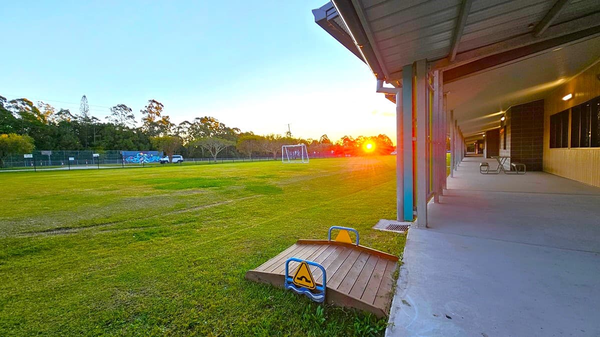 On the left, there are tall trees and bushes bordering a large grassy field. In the middle, there is an expansive lawn with a few scattered concrete utility covers. On the right, there are several buildings with covered walkways and a playground area featuring a red and yellow play structure under a black shade sail. The floor is primarily grass, and the ceiling of the covered walkways is metal with exposed beams. The background includes more trees and additional buildings.