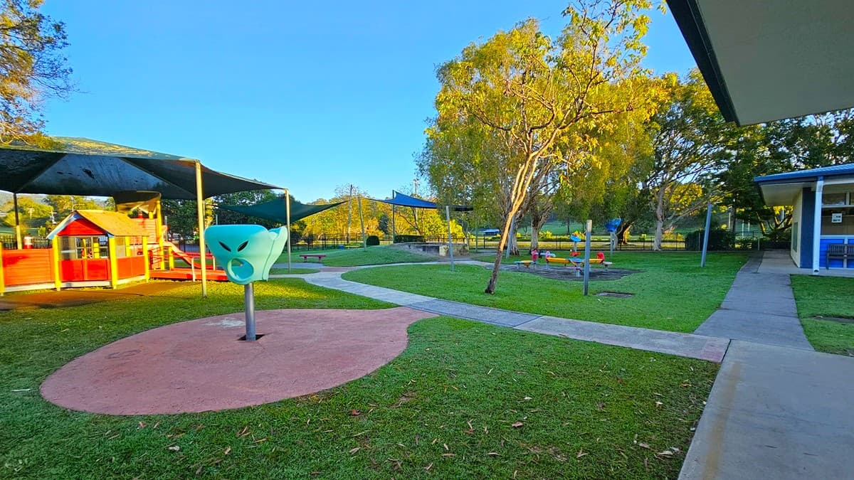 On the left, there is a building with a covered patio area, blue panels, and a white metal fence. In the middle, there is a grassy area with a white metal gate and several red picnic tables in the background. On the right, there is another building with a covered patio, blue panels, and a closed roller door. The floor of the patio areas is concrete, and the ceiling of the patios is white with recessed lighting. The background includes trees and additional buildings.