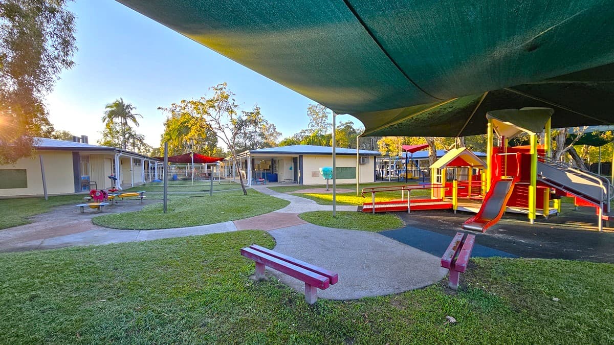 On the left, there is a building with a blue roof and a covered patio area, along with a red picnic table. In the middle, there is a curved concrete path leading to a central grassy area with benches and a playground in the background. On the right, there is a red and yellow play structure with slides and a black shade sail above it. The floor around the play structure is a mix of concrete and rubber matting. The ceiling of the patio areas is white with recessed lighting.