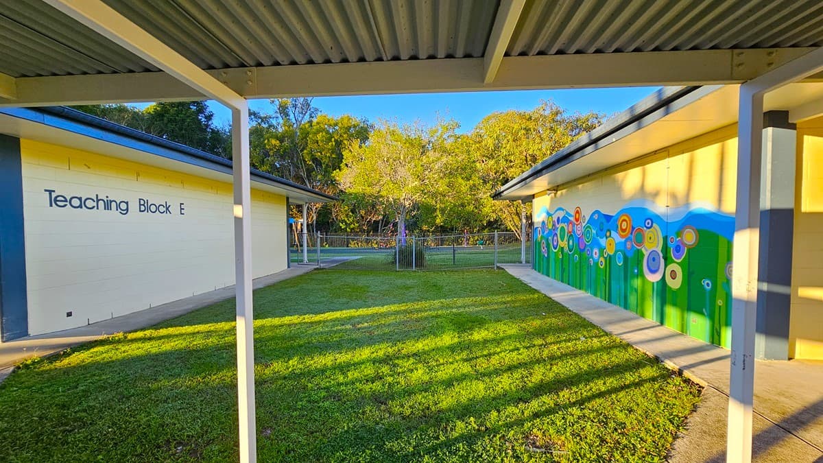 On the left, there is a building labelled "Teaching Block E" with a blue roof and white walls. In the middle, there is a grassy courtyard with a concrete path running along the edges. On the right, there is another building with a colourful mural of flowers on a yellow wall. The floor of the courtyard is grass, and the ceiling of the covered walkway is metal with exposed beams. The background includes trees and a fence.