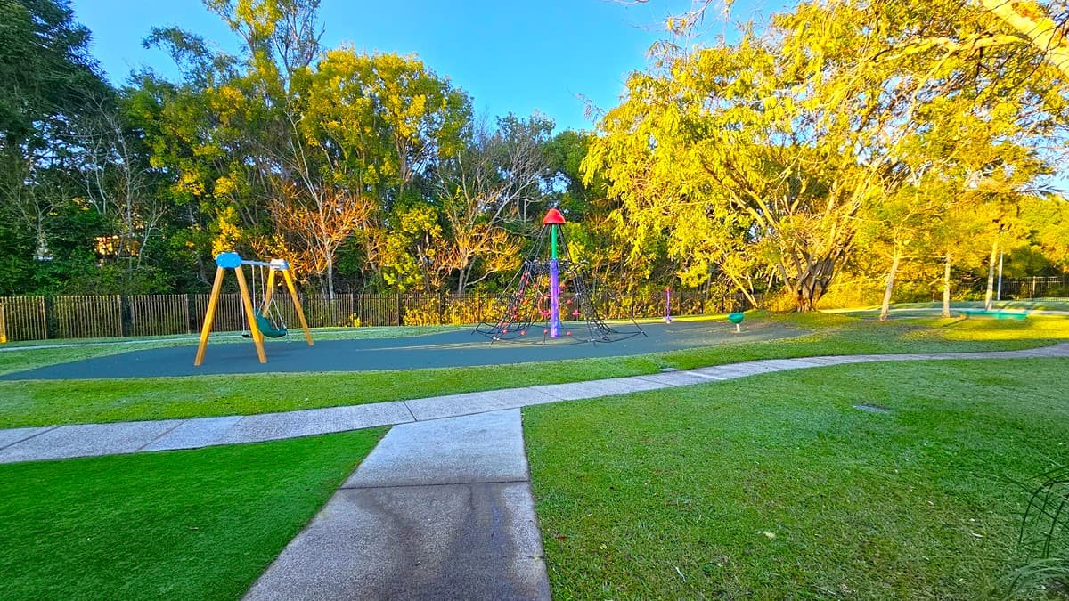 On the left, there is a wooden swing set with two swings on a green rubber mat. In the middle, there is a colourful climbing structure with a red top, also on a green rubber mat. On the right, there are trees with yellow leaves and a grassy area. The floor is a mix of grass and concrete paths. The background includes more trees and a wooden fence.