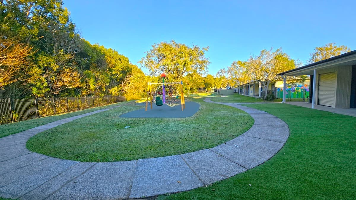 On the left, there is a black metal fence with trees and bushes behind it. In the middle, there is a playground with a wooden swing set on a green rubber mat, surrounded by a curved concrete path. On the right, there is a building with a covered patio area, a roller door, and colourful murals on the walls. The floor is a mix of grass and concrete paths. The background includes more trees and additional buildings.