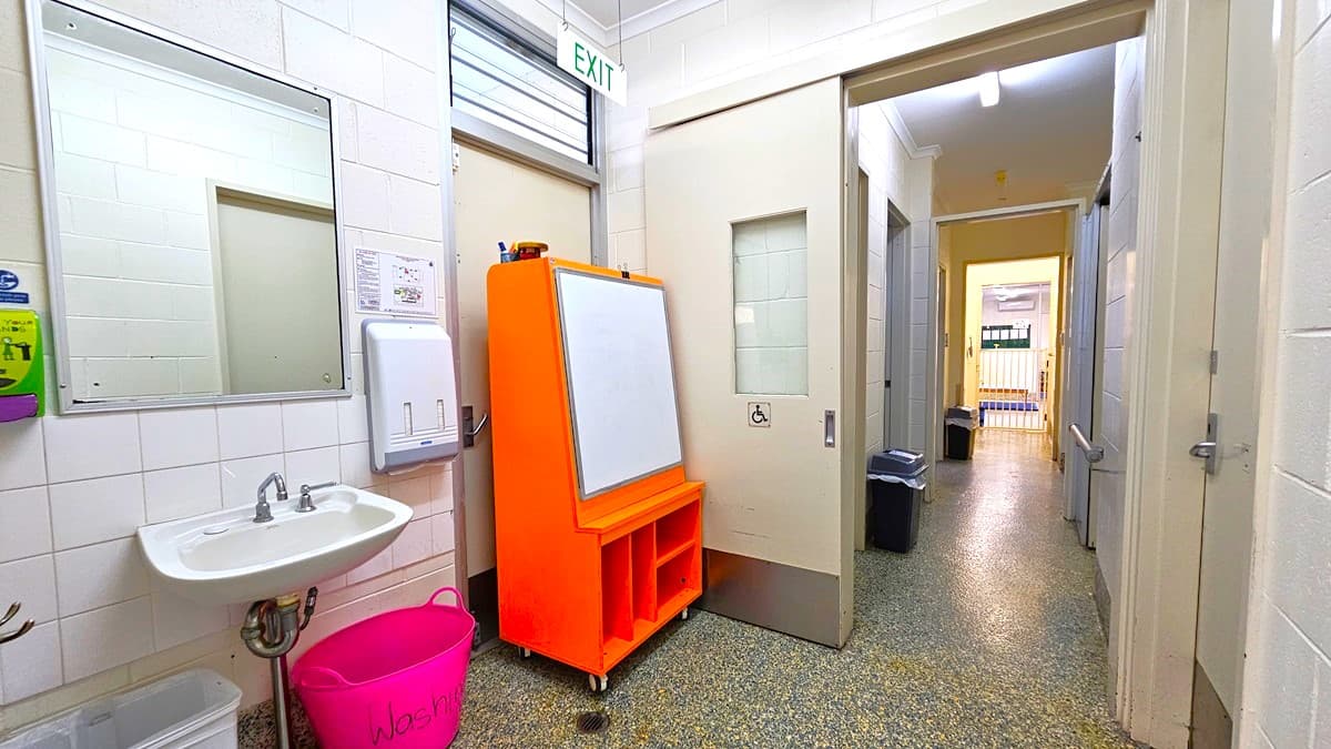 On the left, there is a white tiled wall with a mirror, a sink, a soap dispenser, and a bright pink bin. On the right, there is a corridor with beige walls, a speckled grey floor, and a sliding door with a frosted window. In the middle, there is an orange mobile whiteboard with storage compartments at the bottom. The background features an open doorway leading to another corridor with a gate and a notice board. The ceiling is white with fluorescent lights and an exit sign.