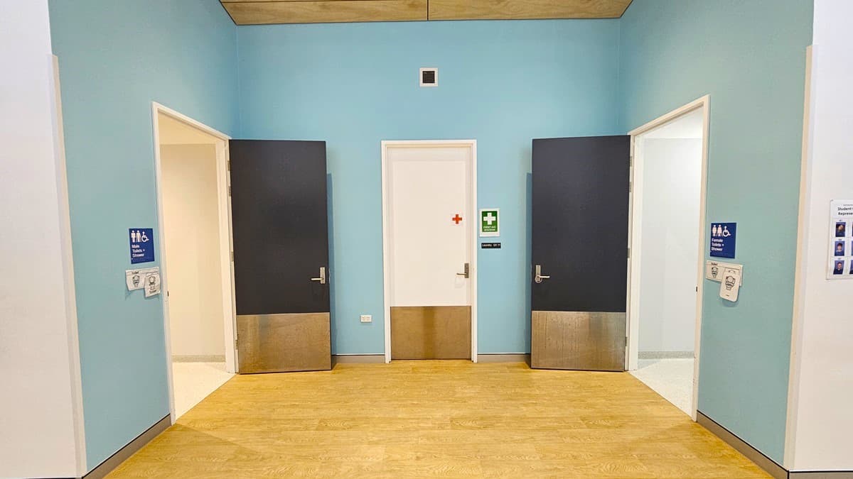 On the left, there is a black door with a silver bottom panel, next to a blue wall with a white door leading to a restroom, marked by a blue sign. In the middle, there is a white door with a red cross and a green first aid sign above it. On the right, there is another black door with a silver bottom panel, next to a blue wall with a white door leading to another restroom, also marked by a blue sign. The floor is light wood, and the ceiling has wooden panels with a small vent in the centre.