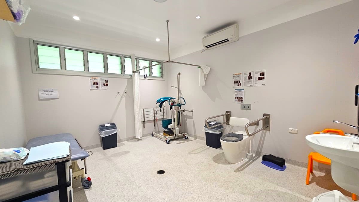 On the left, there is a medical examination table with a blue cushion, a small table with medical supplies, and a black bin. In the middle, there is a patient lift machine and a grey commode chair. On the right, there is a white sink with a soap dispenser, an orange chair, and a white toilet with support bars. The background has a row of frosted windows and several medical posters on the walls. The floor is light grey, and the ceiling has recessed lights and an air conditioning unit.