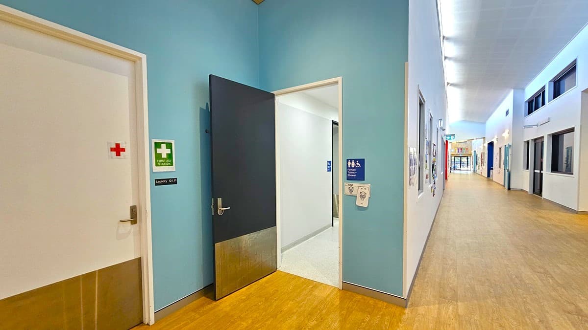 On the left, there is a white door with a red cross and a green first aid sign, next to an open black door leading to a room with white walls and a grey floor. In the middle, there is a blue wall with a sign indicating male toilets and showers, along with an emergency button. On the right, there is a long corridor with white walls, several windows, and a few posters. The floor is light wood, and the ceiling has white tiles with recessed lights and ventilation panels.