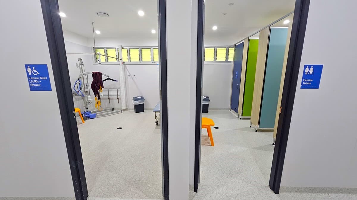 On the left, there is a room with a blue sign indicating female toilets, a shower area with a patient lift machine, a bench, and a black bin. In the middle, there is a wall separating two rooms, with a small orange bench in front. On the right, there is another room with a blue sign indicating female toilets, containing green and blue cubicles, a black bin, and a white wall. The floor is light grey, and the ceiling has recessed lights and ventilation panels.