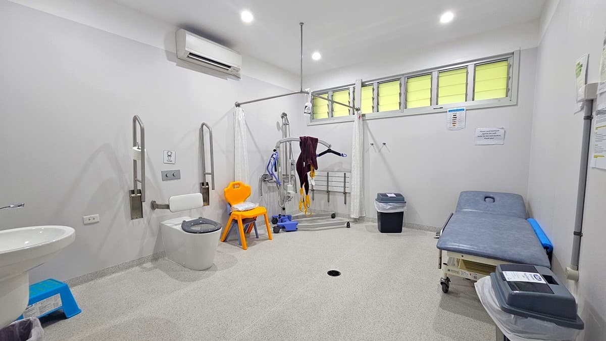On the left, there is a white sink with a soap dispenser, a white toilet with support bars, and a blue box on the floor. In the middle, there is a patient lift machine with a harness, an orange chair, and a black bin. On the right, there is a grey examination table with a blue cushion, a small table with medical supplies, and a white wall with a few notices. The background has a row of frosted windows. The floor is light grey, and the ceiling has recessed lights and an air conditioning unit.