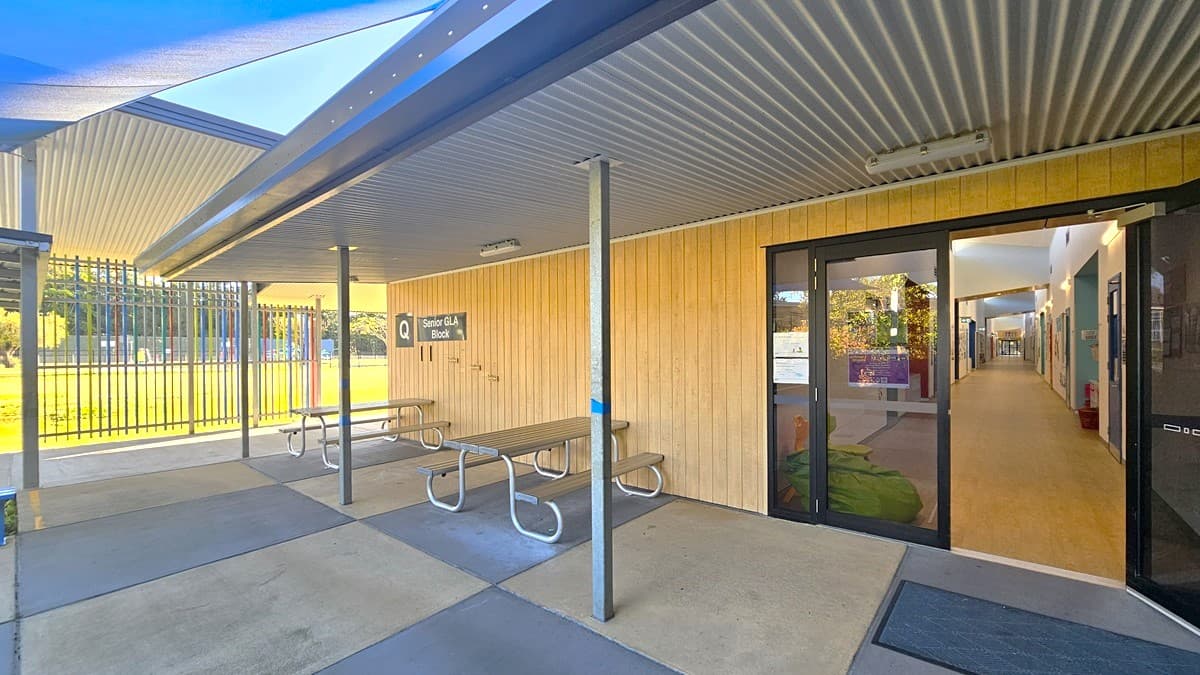 On the left, there are metal picnic tables under a covered area with a corrugated metal roof supported by grey poles. In the middle, there is a beige wooden wall with a sign reading "Senior Unit Block 1" and a glass door leading inside. On the right, there is a glass entrance door showing an interior hallway with lights and decorations. The floor is a mix of concrete outside and light-coloured flooring inside.