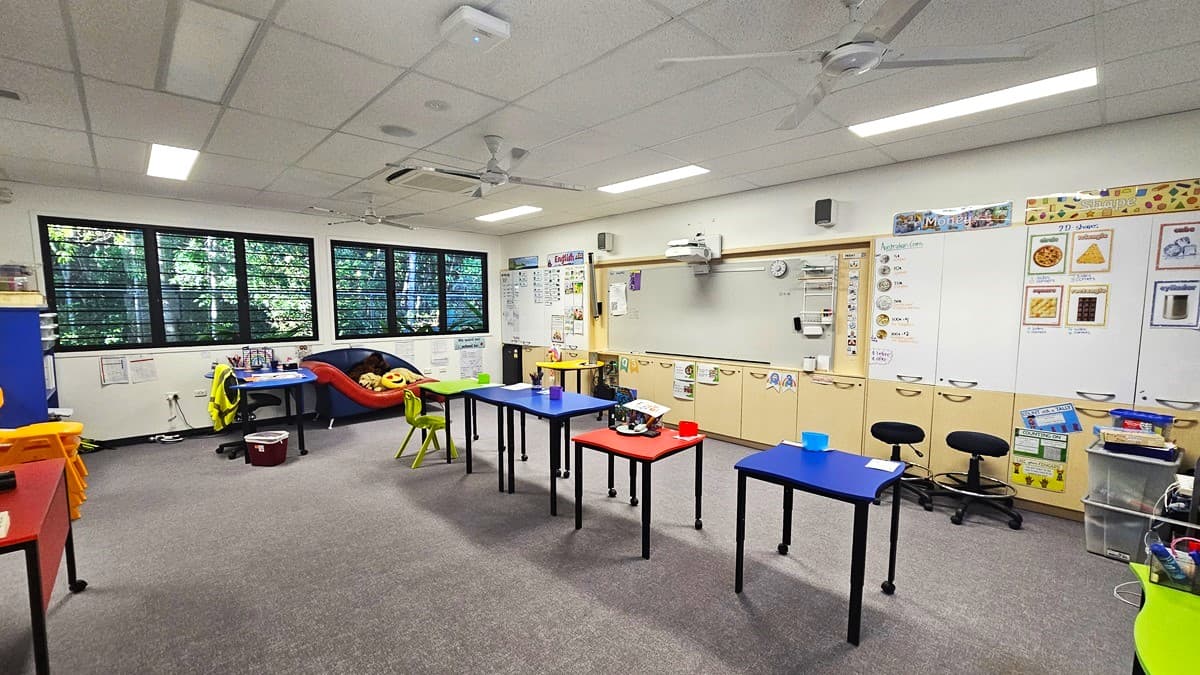 On the left, there are large windows with black frames, showing greenery outside. Below the windows, there is a red cushioned reading nook with a small table and chairs. In the middle, there are several tables in blue and red with various educational materials on them. On the right, there are white cabinets and bulletin boards displaying educational posters and artwork. The floor is carpeted in grey, and the ceiling is white with recessed lighting and ceiling fans.