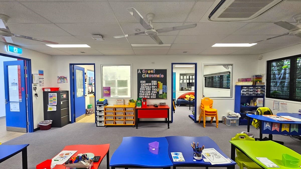 On the left, there is a blue door with a window, a recycling bin, and a whiteboard. In the middle, there is a red table with yellow drawers underneath, a bulletin board with "A Great Classroom" written on it, and a doorway leading to another room with a round table and chairs. On the right, there are shelves with educational materials, a yellow chair, and large windows with black frames. The floor is grey carpet, and the ceiling is white with recessed lighting, and ceiling fans.