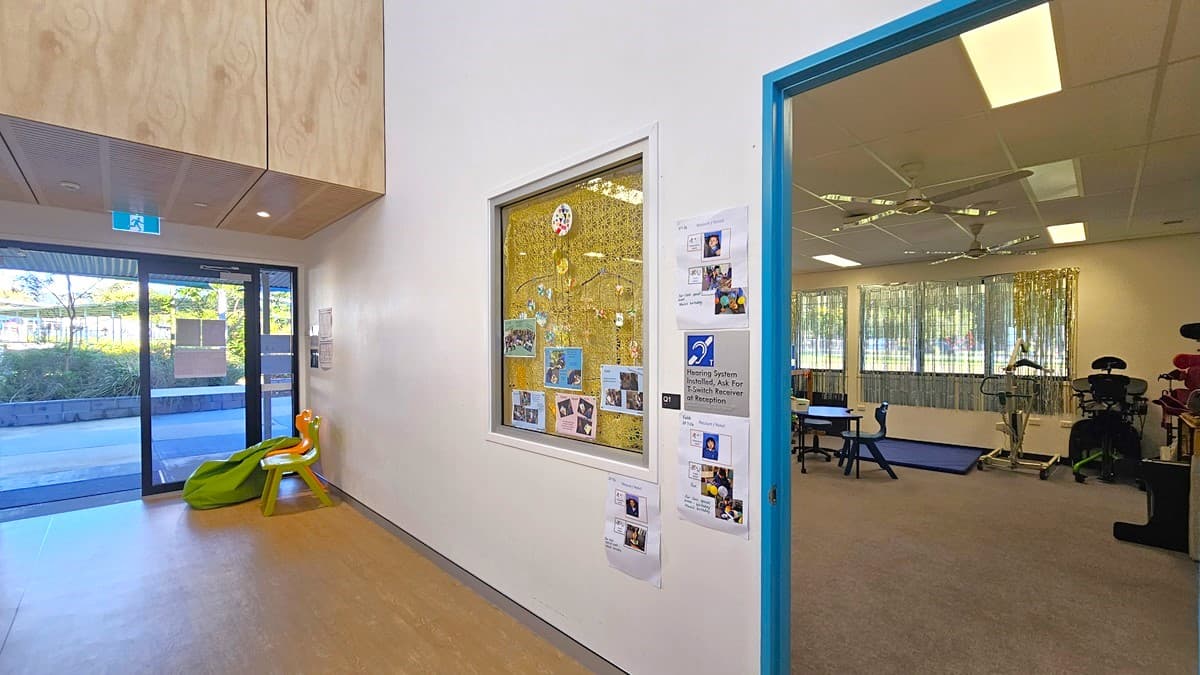 On the left, there is a glass door with a blue frame leading outside, next to a green and orange slide. In the middle, there is a large window displaying children's artwork and photos, with more pictures and notices pinned to the wall below it. On the right, there is an open doorway with a blue frame leading to a room with exercise equipment, tables, and chairs. The floor is light-coloured wood, and the ceiling is white with recessed lighting and ceiling fans.