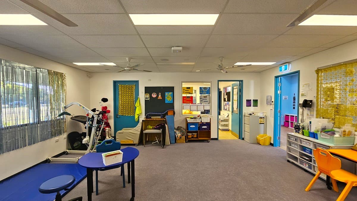 On the left, there is exercise equipment next to a window with vertical blinds. In the middle, there is a blue table with a small sign on it, surrounded by blue chairs, and a doorway leading to another room with shelves and storage units. On the right, there are blue-framed doors, a yellow bin, and white shelves with various supplies. The floor is grey carpet, and the ceiling is white with recessed lighting and ceiling fans.