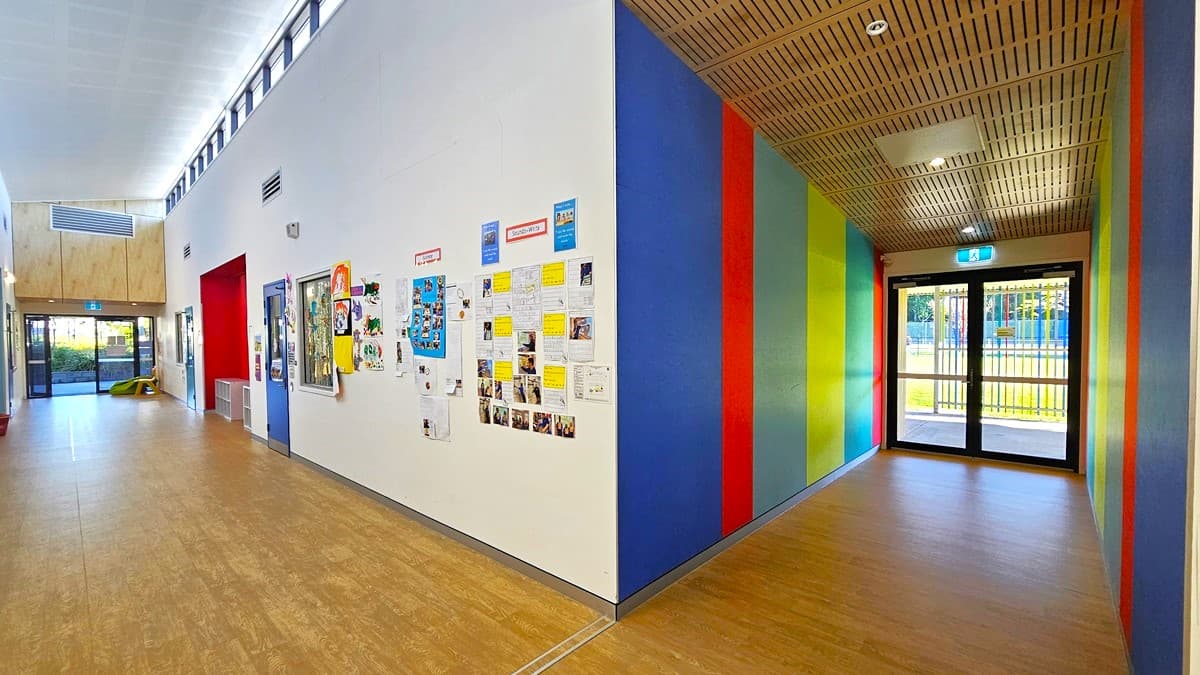 On the left, there is a long hallway with light wooden flooring, white walls, and a series of windows near the ceiling. Bulletin boards with various notices and pictures are mounted on the wall. In the middle, there is a corner where the hallway turns, leading to a colourful corridor with vertical panels in blue, red, green, and yellow. On the right, the colourful corridor has a glass door at the end, leading outside. The ceiling is white with recessed lighting and wooden panels.