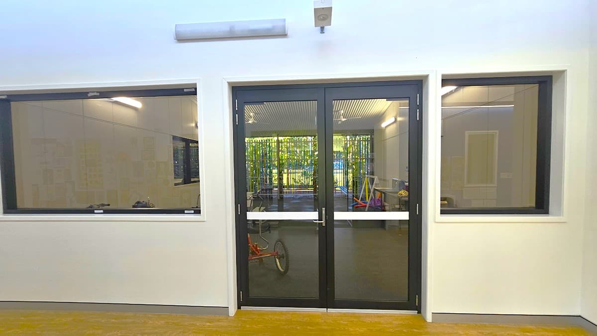 On the left, there is a large window with a black frame, showing an interior room with white walls and a black floor. In the middle, there are double glass doors with black frames, leading to a room with exercise equipment and a view of greenery outside. On the right, there is another large window with a black frame, similar to the one on the left. The floor is light-coloured wood, and the ceiling is white with a linear light fixture and a security camera.