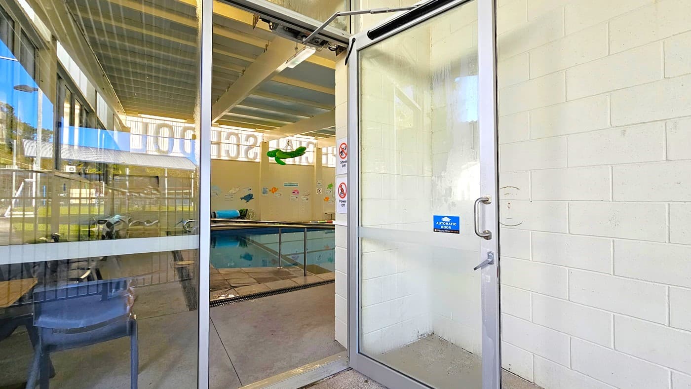 On the left, there is a glass wall with a sliding door, a grey chair, and a view of an indoor pool area with a green slide. In the middle, there is a glass door with a blue sign and a metal handle, leading to the pool area. On the right, there is a white brick wall with a no smoking sign. The floor is light grey concrete, and the ceiling has exposed beams and fluorescent lights. The background includes the pool, poolside chairs, and windows letting in natural light.