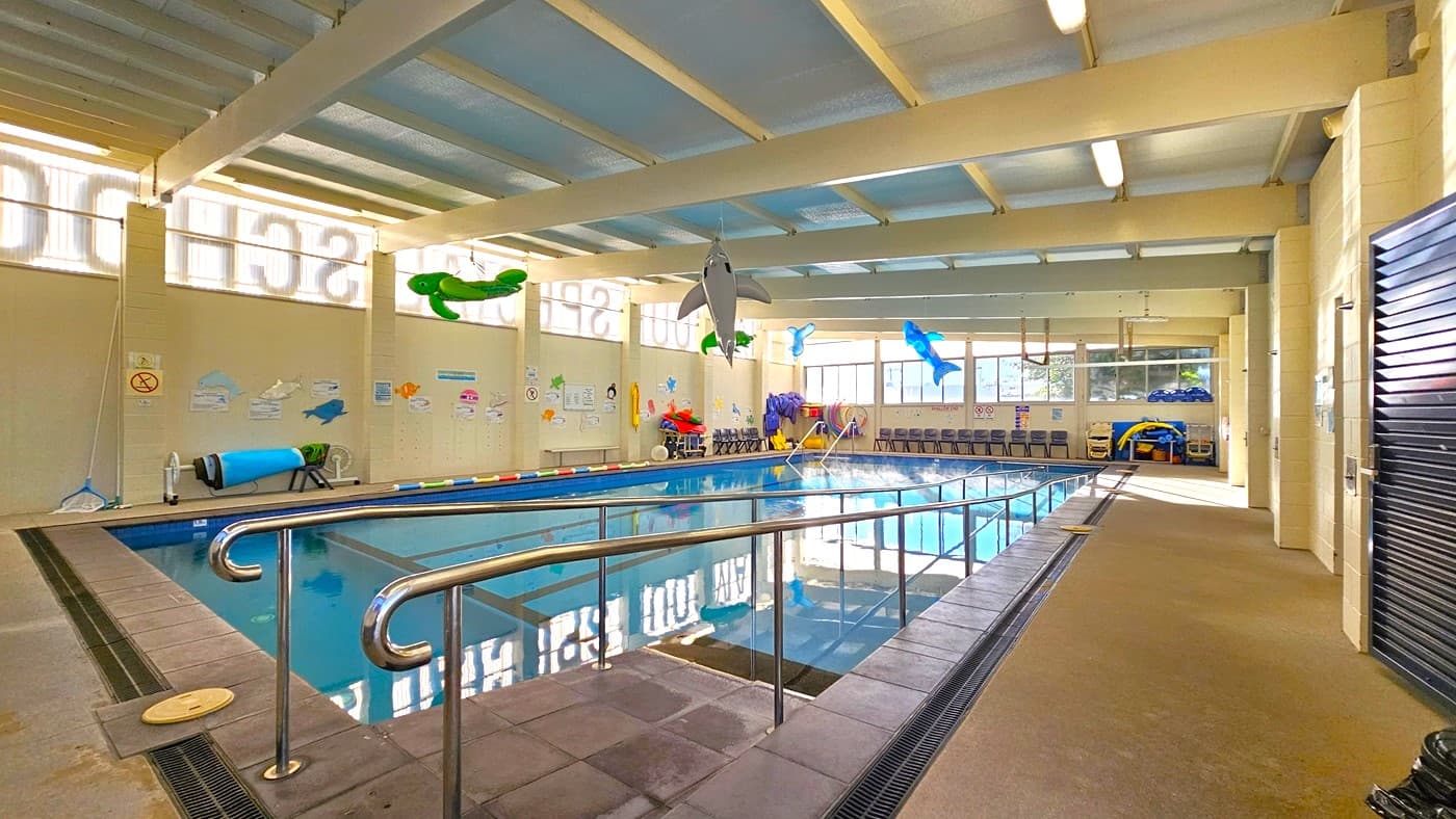 On the left, there is a tiled pool deck with a metal handrail leading into the water, colourful wall decorations, and a no diving sign. In the middle, there is a large indoor swimming pool with lane markers and floating toys, including a green slide and a dolphin. On the right, there are rows of chairs along the wall, a small play area with toys, and a black door. The floor is a mix of tiles and concrete, and the ceiling has exposed beams with skylights and fluorescent lights.