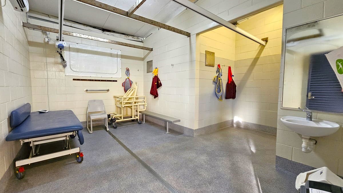 On the left, there is a blue cushioned examination table on wheels, a white grab bar, and a yellow patient lift machine next to a bench. In the middle, there are hooks on the wall with hanging red and yellow towels. On the right, there is a white sink with a mirror above it and a soap dispenser. The floor is grey with a drainage channel, and the ceiling has exposed beams and fluorescent lights. The background includes a frosted window and a ventilation grate.