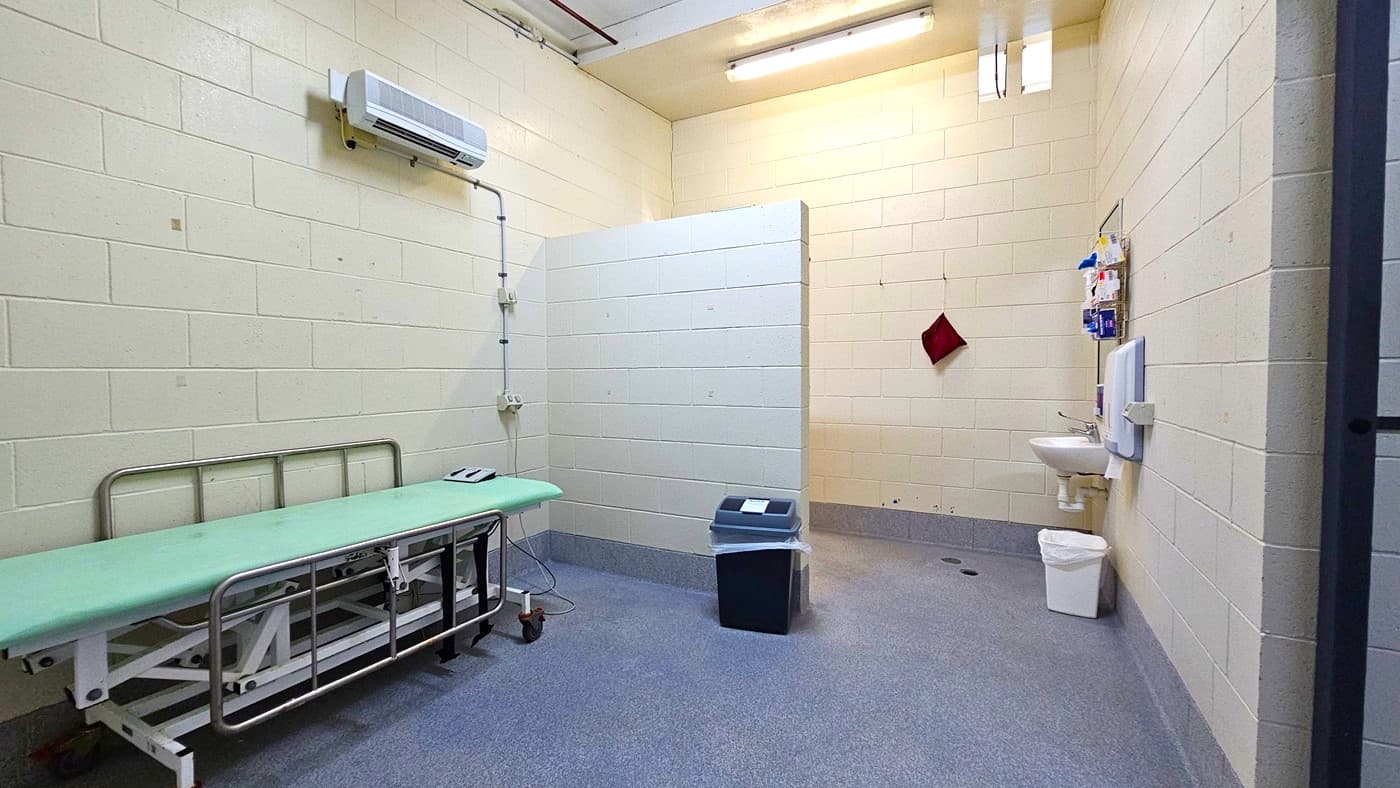 On the left, there is a green cushioned examination table on wheels with a control panel, and an air conditioning unit mounted on the wall above it. In the middle, there is a partial wall divider and a blue bin on the floor. On the right, there is a white sink with a mirror, a soap dispenser, a red towel hanging on a hook, and a white bin below. The floor is grey with a drainage hole, and the ceiling has exposed pipes and fluorescent lights. The background includes cream-coloured brick walls.