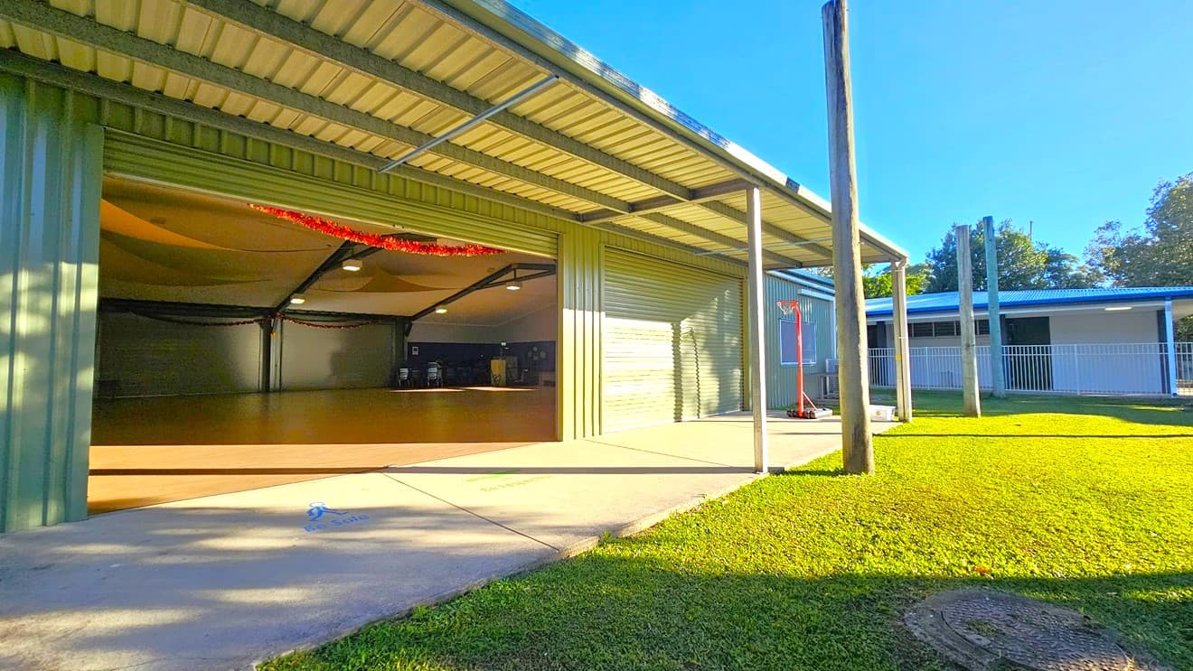 On the left, there is a large open garage with a corrugated metal roof and walls, revealing an empty interior with a wooden floor. In the middle, there is a concrete path leading from the garage to the outside. On the right, there is a grassy area with a basketball hoop and a building with a blue roof and white railings. The background includes trees and additional grassy areas. The floor is a mix of concrete and grass, and the ceiling of the garage is corrugated metal.