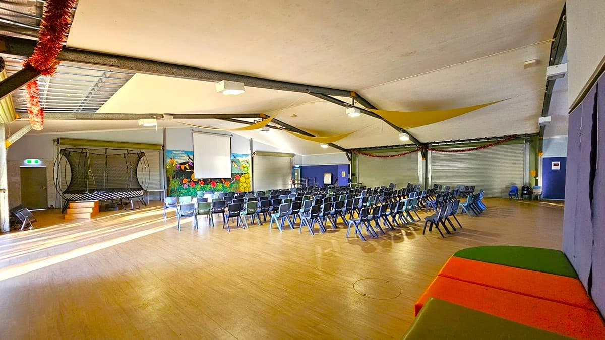 On the left, there is a trampoline with a safety net and a small stage area with a blue door. In the middle, there are rows of black chairs facing a projector screen and a colourful mural of a garden on the back wall. On the right, there are two large roller doors and a blue wall with a few chairs stacked against it. The floor is wooden, and the ceiling is white with exposed metal beams and yellow fabric draped across, along with fluorescent light fixtures.