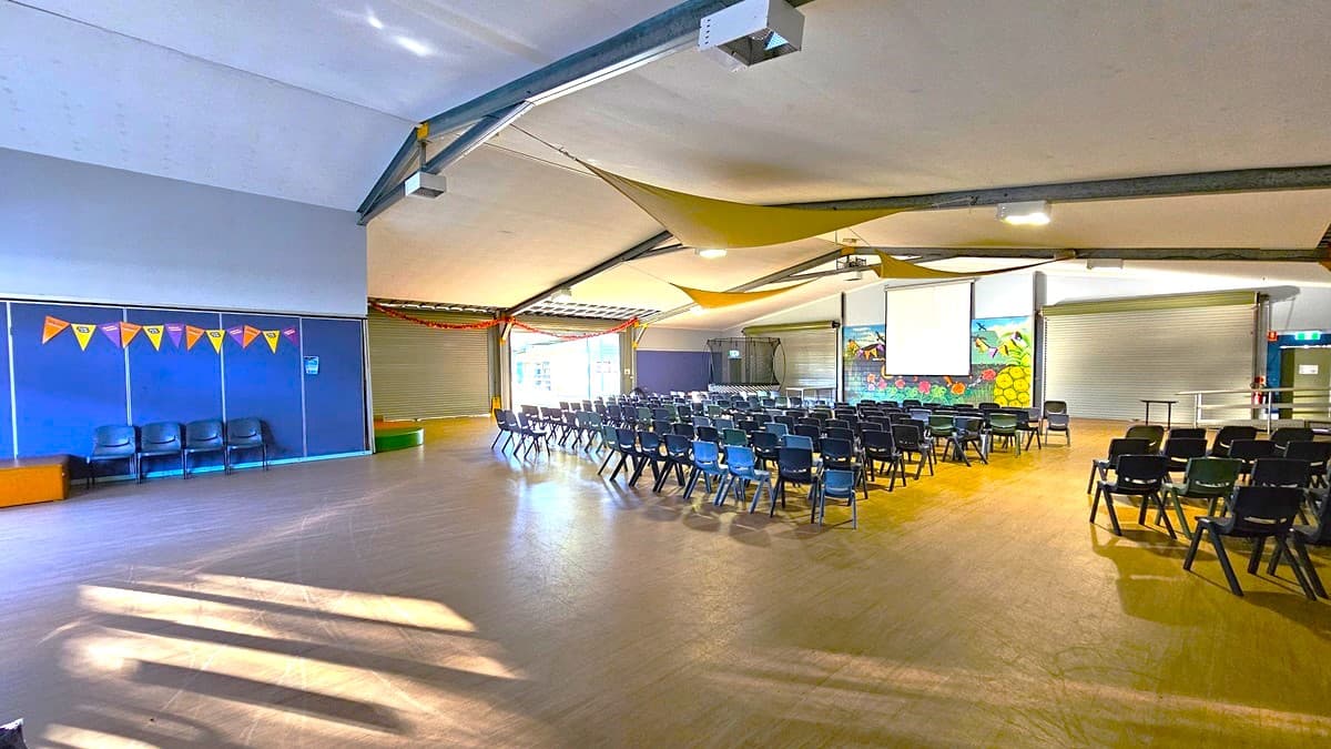 On the left, there is a trampoline with a safety net and a small stage area with a blue door. In the middle, there are rows of black chairs facing a projector screen and a colourful mural of a garden on the back wall. On the right, there are two large roller doors and a blue wall with a few chairs stacked against it. The floor is wooden, and the ceiling is white with exposed metal beams and yellow fabric draped across, along with fluorescent light fixtures.