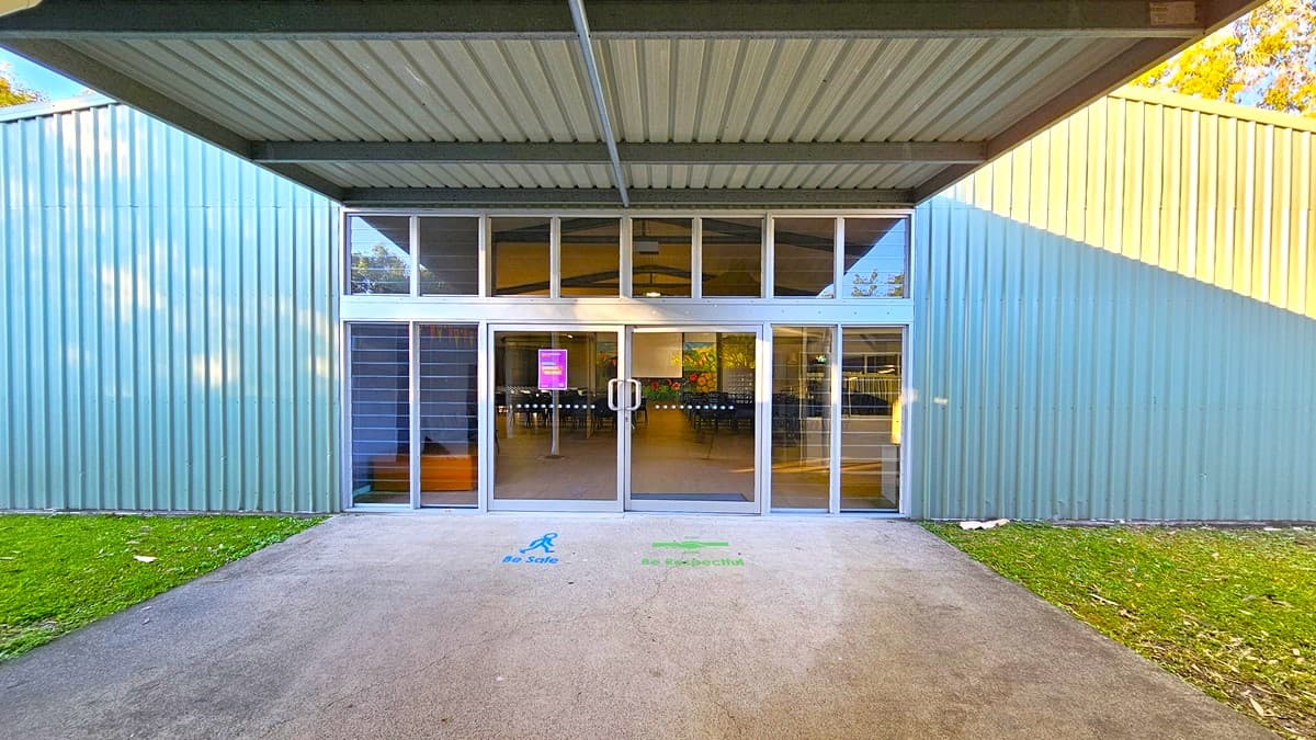 On the left, there is a green metal wall with vertical ridges and a small patch of grass. In the middle, there is a large glass entrance with double doors, leading into a building with visible rows of chairs and a colourful mural inside. On the right, there is another green metal wall with vertical ridges and a small patch of grass. The floor is a concrete path leading up to the entrance, and the ceiling is a corrugated metal roof extending over the entrance area.