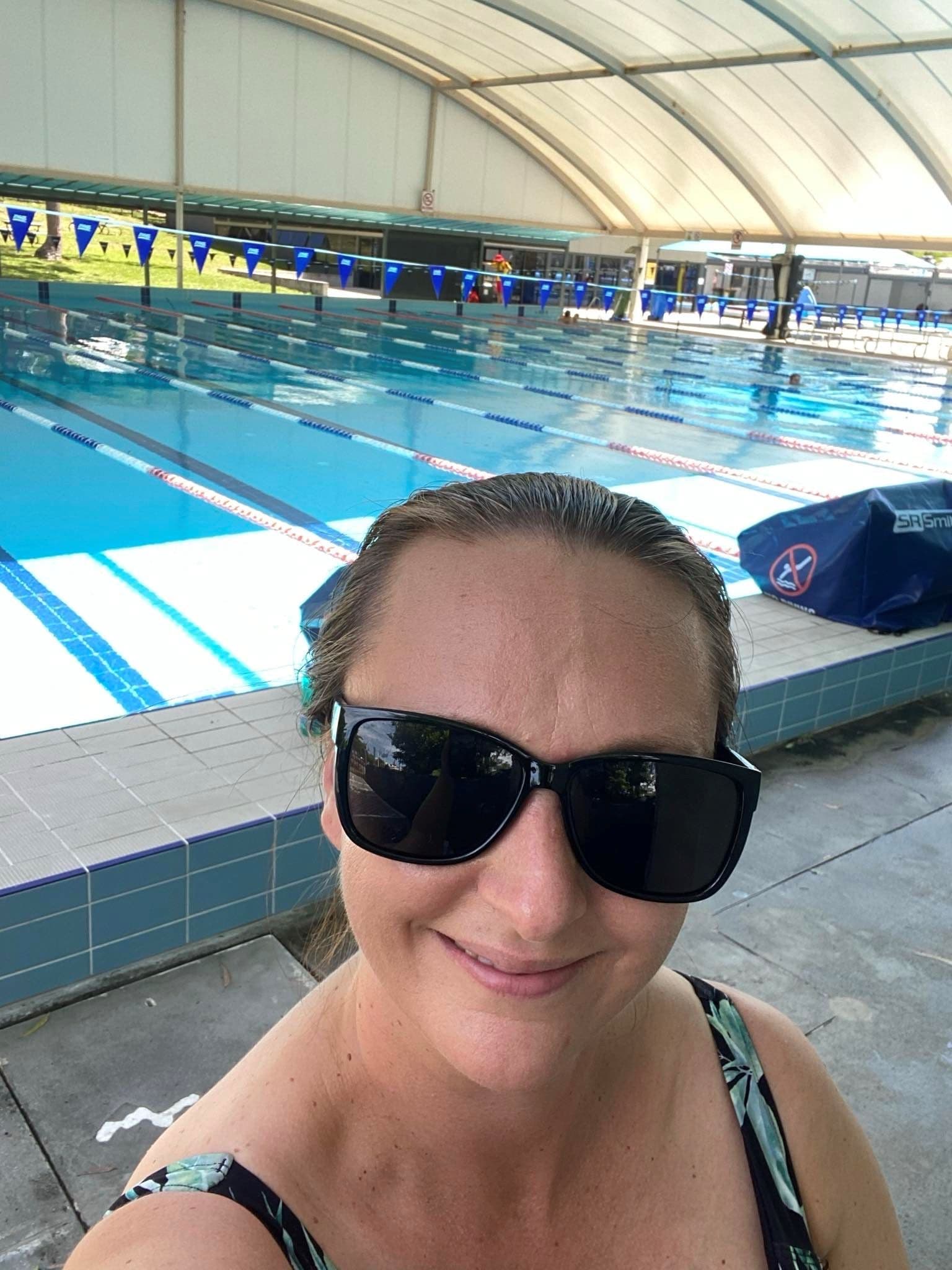 Person wearing sunglasses by a swimming pool, smiling at the camera.