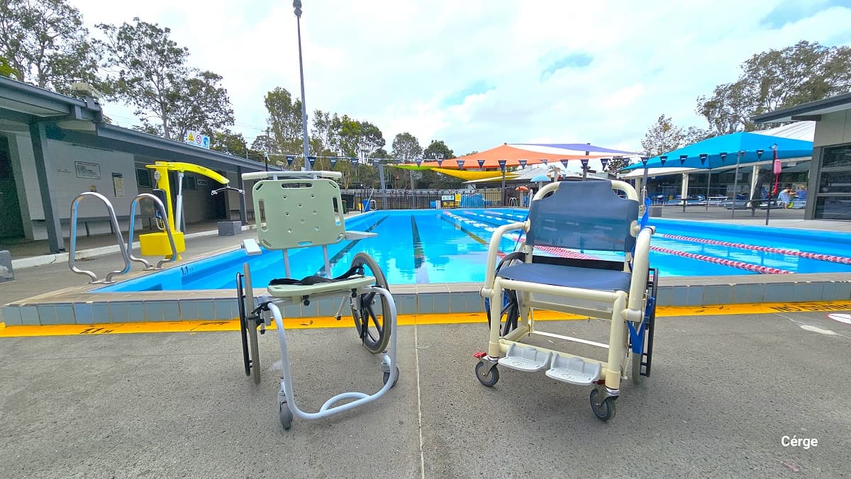 Two aquatic wheelchairs on the pool deck of the 25m, 4-lane outdoor pool. The left aquatic wheelchair has a light green seat and a back support. It has a light blue frame for the foot and a light blue handle for pushing. The right wheelchair has a waterproof cloth cover for seat and back support. It has two-foot support and is composed of cream metal frames.