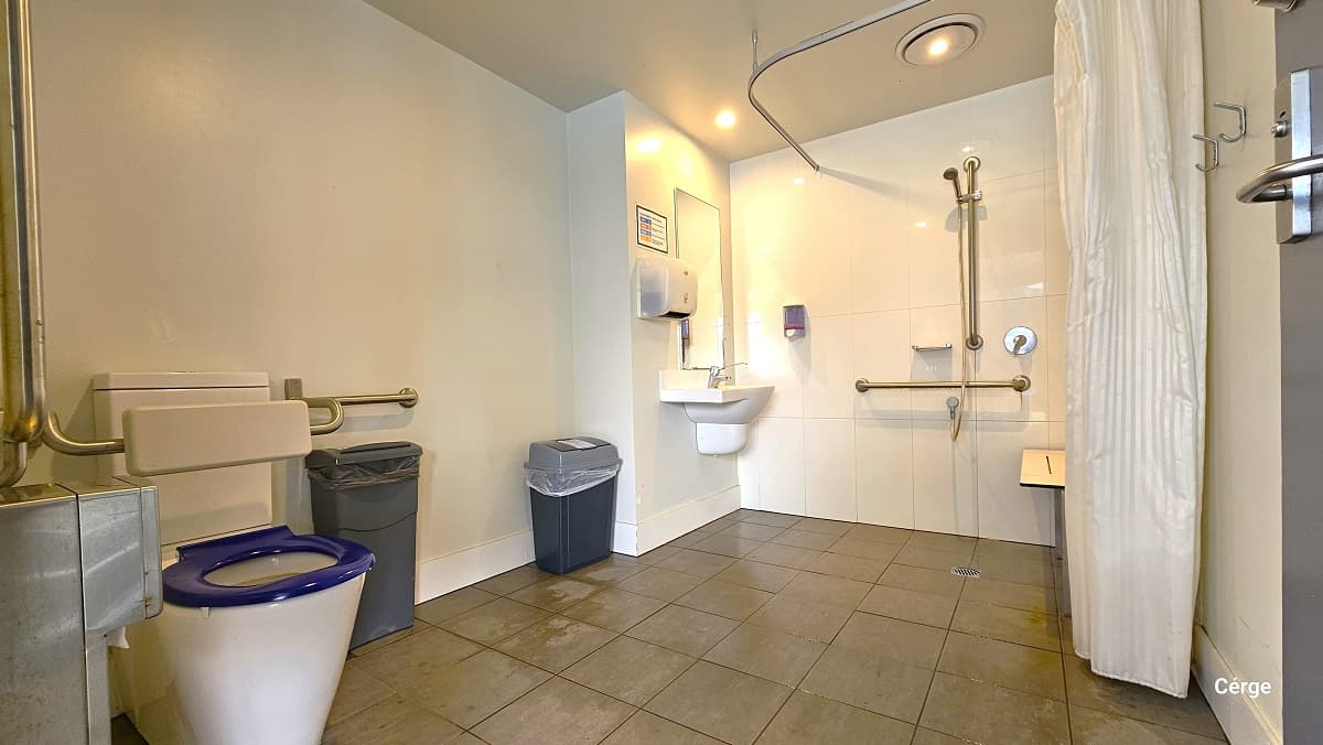 Inside the accessible bathroom at Nerang Aquatic Centre. Near the door is the toilet with a blue toilet cover and white back support. Grab bars are available on the walls near the toilet. On the left of the toilet is the tissue dispenser. There are two grey garbage bins inside the toilet and shower area. The shower has a tall white shower curtain, a wall-mounted shower seat, a shower head with grab bars, and a small basin with a tall wall mirror. The bathroom has a tiled floor with white walls.