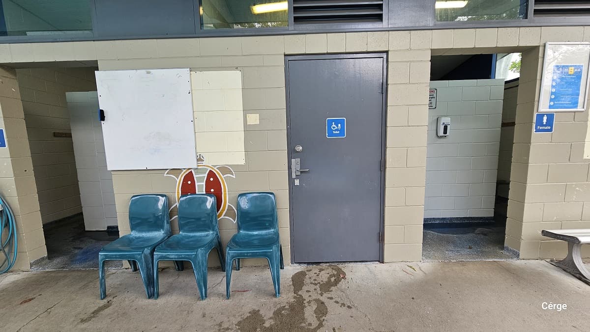 Accessible toilet and a female toilet at Nerang Aquatic Centre. It is marked by a small blue sign and dark grey doors. Three blue-green plastic chairs are outside, on the left side of the accessible toilet. There is an open corridor to the left. The toilet inside has a white brick wall, while outside is a light brown brick wall with a small glass window and an air ventilation window. Below the white bulletin is a colorful graphic of a turtle.