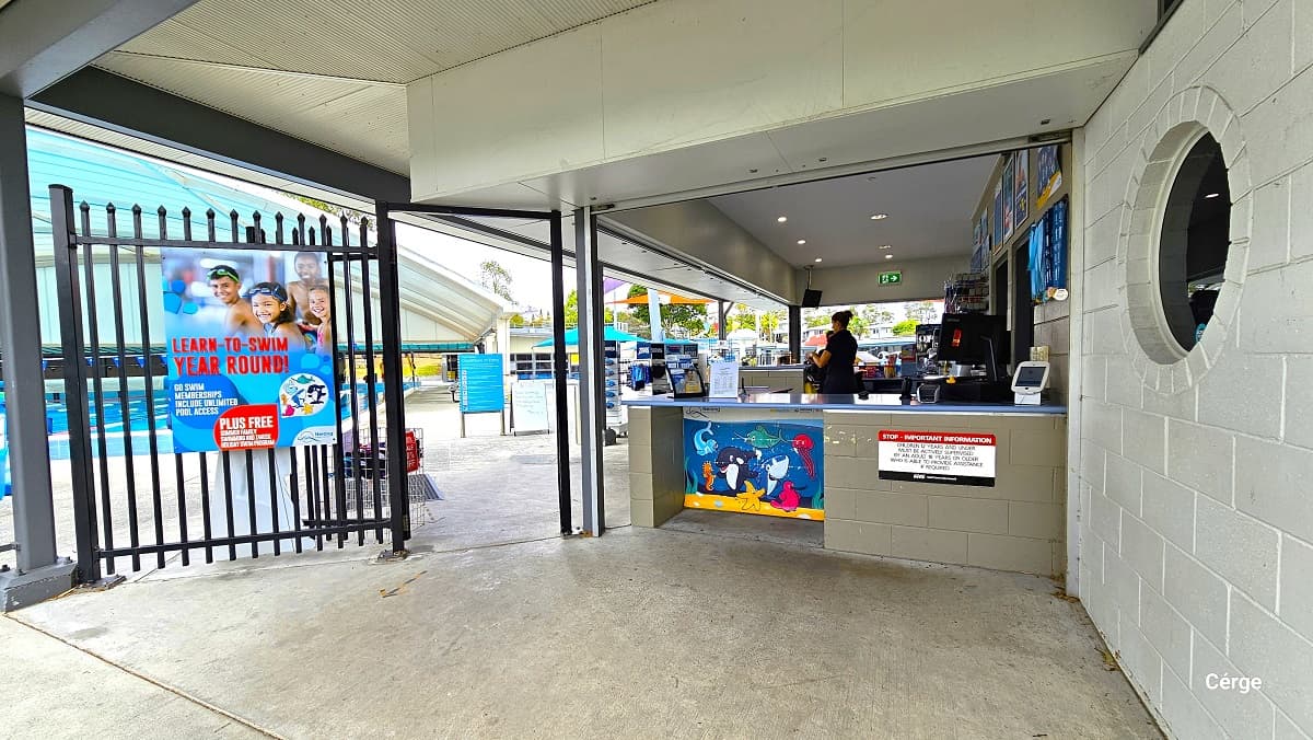 A low-height reception counter adjoining a tall black pool fence creates an entry point to pay or scan for admittance to the Nerang Aquatic Centre. There is a large dark blue signboard containing the opening hours, the different facilities, and their rules and conditions and a red bell-shaped lamp post near the black fence. On the right is a small, black, open sign and a garden filled with shrubs and trees with abundant foliage.