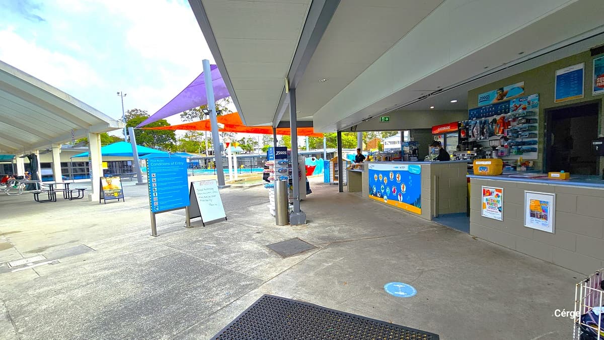 There is a small retail area with aquatic items, snacks, and beverages after entering through the gate and admittance through the reception desk. It has a grey brick reception desk with a light blue countertop designed with colorful animated ocean creatures. Directly in front is the blue sign board for the conditions of entry to the pool and a small whiteboard. From here, guests can see the different outdoor aquatic amenities.