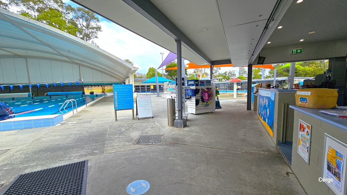 View standing from the retail area in Nerang Aquatic Centre. To the left is the main 10-lane, 25m pool with a white dome structure. The pool has red, blue, and white floating rope lanes, a string of triangular blue flags overhang, and a pool ladder distinguished by the curvy handle. The start of the pool has an elevated blue and white tiled floor and diving blocks on top with a blue cover. Directly ahead is a small splash park and toddler wading pool.