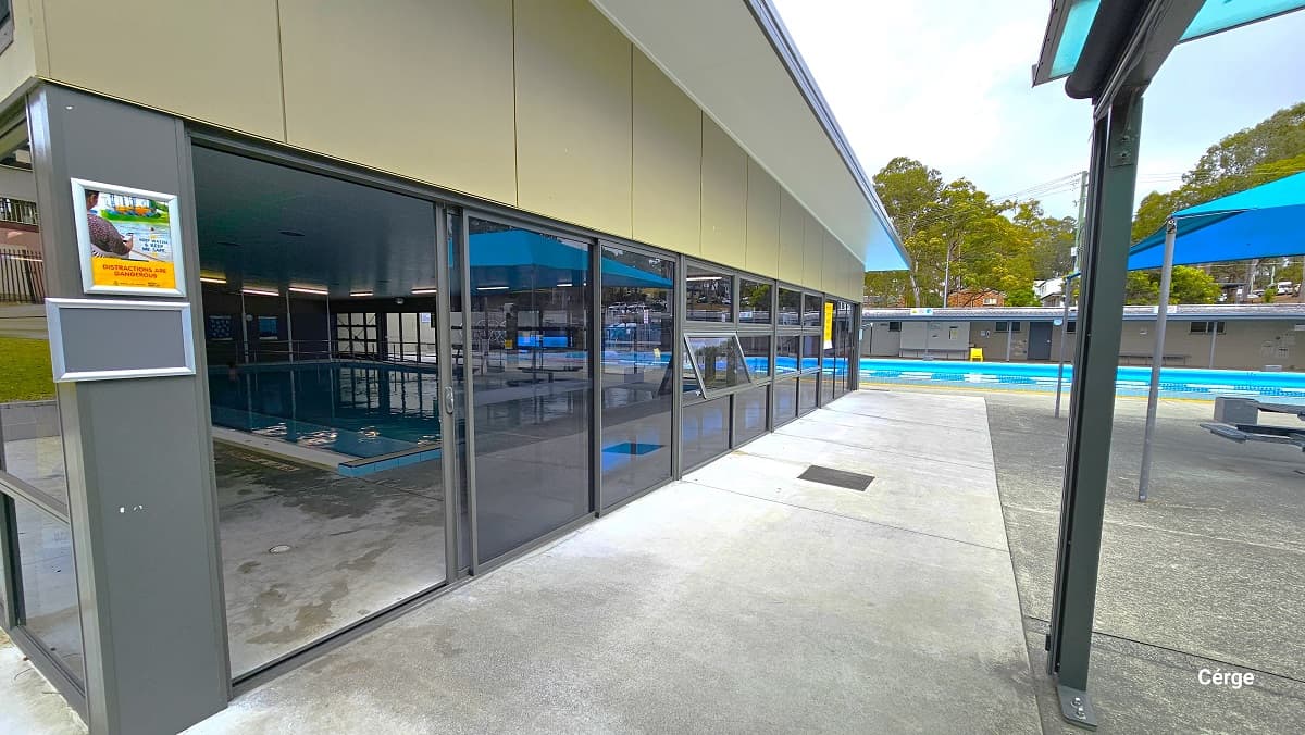 Low-set grey building with tinted glass walls and sheet metal roofing of the indoor programs pool at Nerang Aquatic Centre. It has a cream-colored wall and a white ceiling. On the left grey foundation support are two silver frames with graphics. The indoor pool is behind the large blue tent with a grey picnic table.
