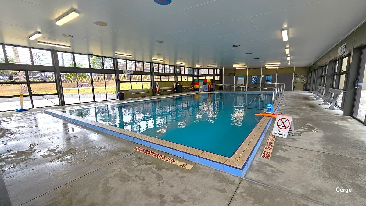 Near the pool edge is a warning sign for shallow water of 0.9 m painted on the floor. There is also a small white sign board for the No Diving warning. On both sides of the pool deck are the benches placed against the wall. On the grey wall to the back, divided by white lines, are adverts. There is a small section on the left wall to the back with colorful cubes.