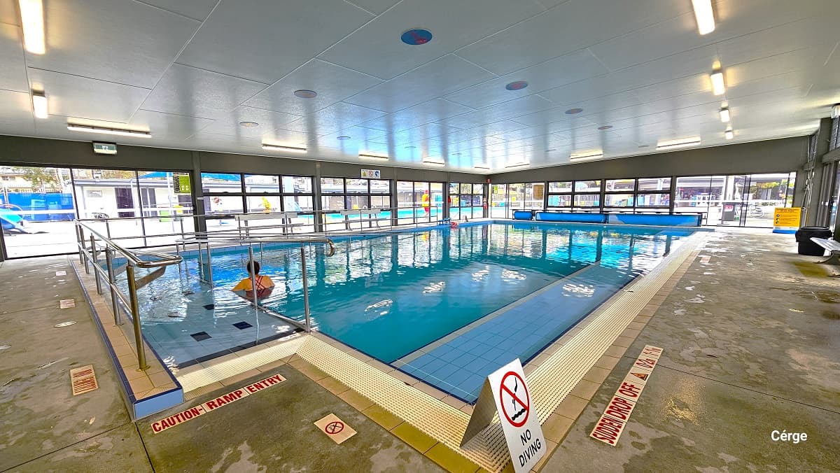 View from the ramp access of the indoor programs pool. At the entry is a red letter warning for “Caution - Ramp Entry.” A no entry sign is placed on the pool deck directly in front of the ramp access with handrails. Near the No Diving white sign board is another caution for “Sudden drop off” detailed on the right pool deck. Further to the back is a black garbage bin. There are blue circles on the ceiling.