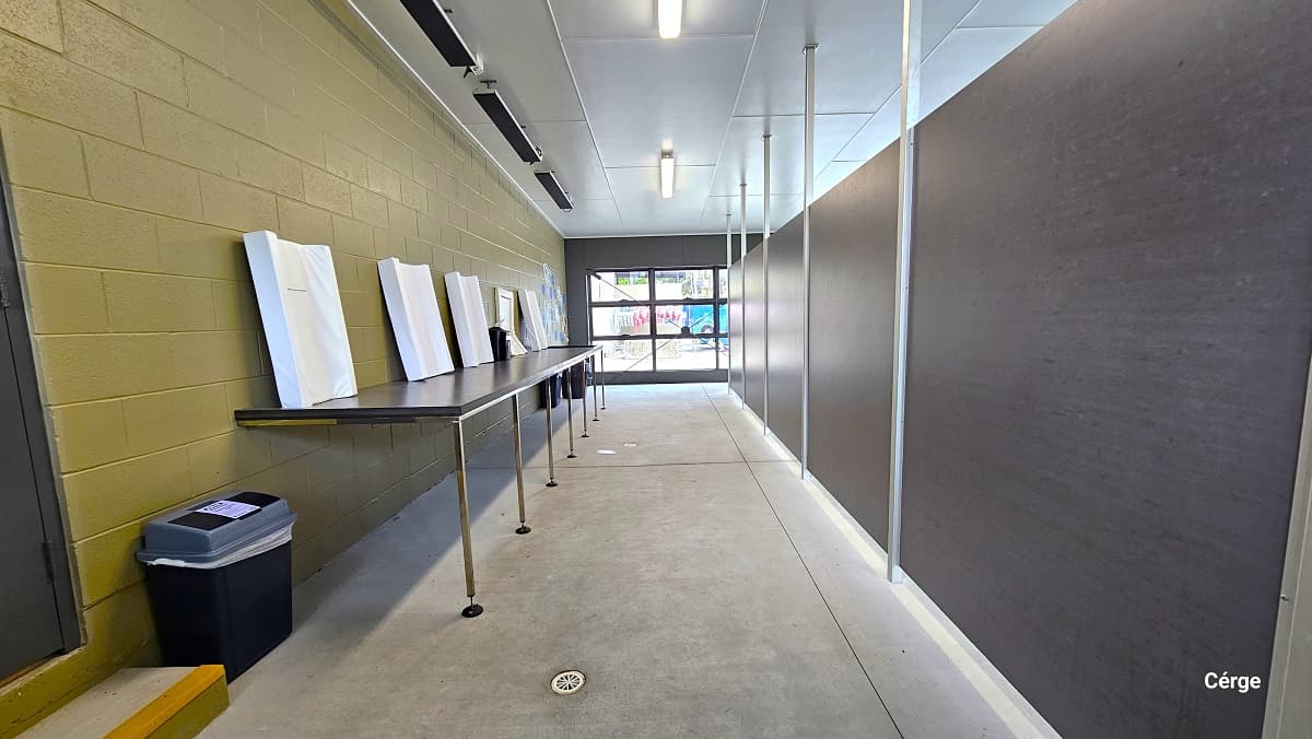 Behind the grey back wall, separated by metal frames connected from the ceiling to the floor. There is a long brown table with white frames on top of it. The table is mounted on the gold brick wall to the left. The cemented floor is smooth, with a white hole for water draining. There is a single-step stair in front of a dark grey door to the left. There are also dark grey rectangular shapes on the ceiling near the edge.