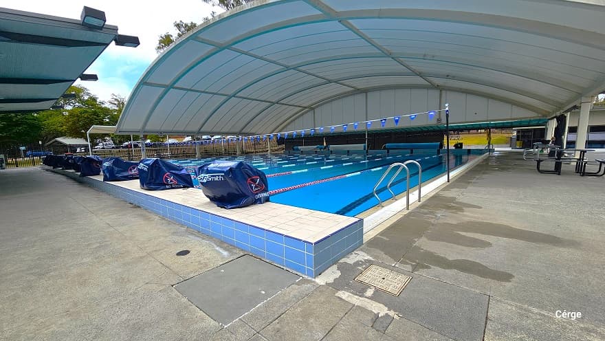 The 25m, 10 lane pool with a large white shaded doom structure at the outdoor aquatic center in Nerang Aquatic. There are two strings of triangular blue flags wrapped around the black poles on the opposite side of the pool. Diving blocks with safety cover are placed on each lane, separated by red, blue, and white floating rope lanes. It has a wide pool deck with three long rolls of blue waterproof tarpaulin at the back.