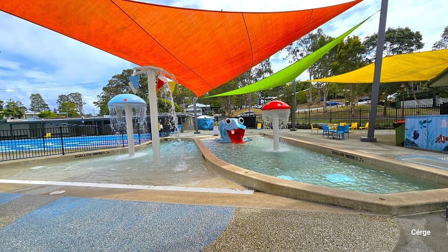 There are red, light green, and yellow shade sails protecting the splash park, toddler wading pool, and the seating area. There is a red mushroom fountain, a large blue frog with a short red water slide, a light blue mushroom fountain, and a cone bucket area with red, blue, and yellow colors. The seating section to the far right has yellow and light blue plastic chairs and tables.