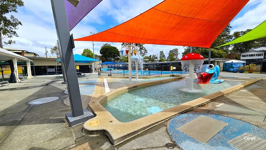 The image shows the Nerang Aquatic Centre's outdoor pool area, featuring a 25m, 10-lane pool covered by a white dome on the left side. There are red and light green shade sails protecting the splash park and toddler wading pool. Water features include a red mushroom fountain, a large blue frog water slide, a light blue mushroom fountain, and a cone bucket area with red, blue, and yellow colors. At the back, there are two large blue tents and a 25m, 4-lane pool separated by a black fence.