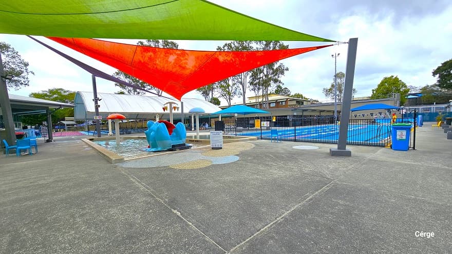 View near the seating section next to the Splash park and Toddler wading pool. The red, light green and purple shade sails are wrapped around the large metal rectangular poles. Splash Park and the Toddler wading pool are separated from the 25m, 4-lane pool by a low black fence. It also has a blue triangular flag, floating rope lanes, and pool ladders. Around the pool deck are yellow plastic chairs.