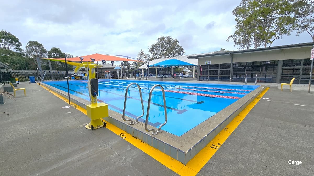 25m, 4-lane pool at Nerang Aquatic Centre. A yellow line is painted around the pool with a 1.1m label indicating the depth of the pool. There is a yellow pool hoist on the left side of the pool. Several pool ladders are placed all around the pool sides. Yellow waterproof plastic chairs are available on the pool deck. There is a beige, yellow, and light green shade sails at the end of the pool.
