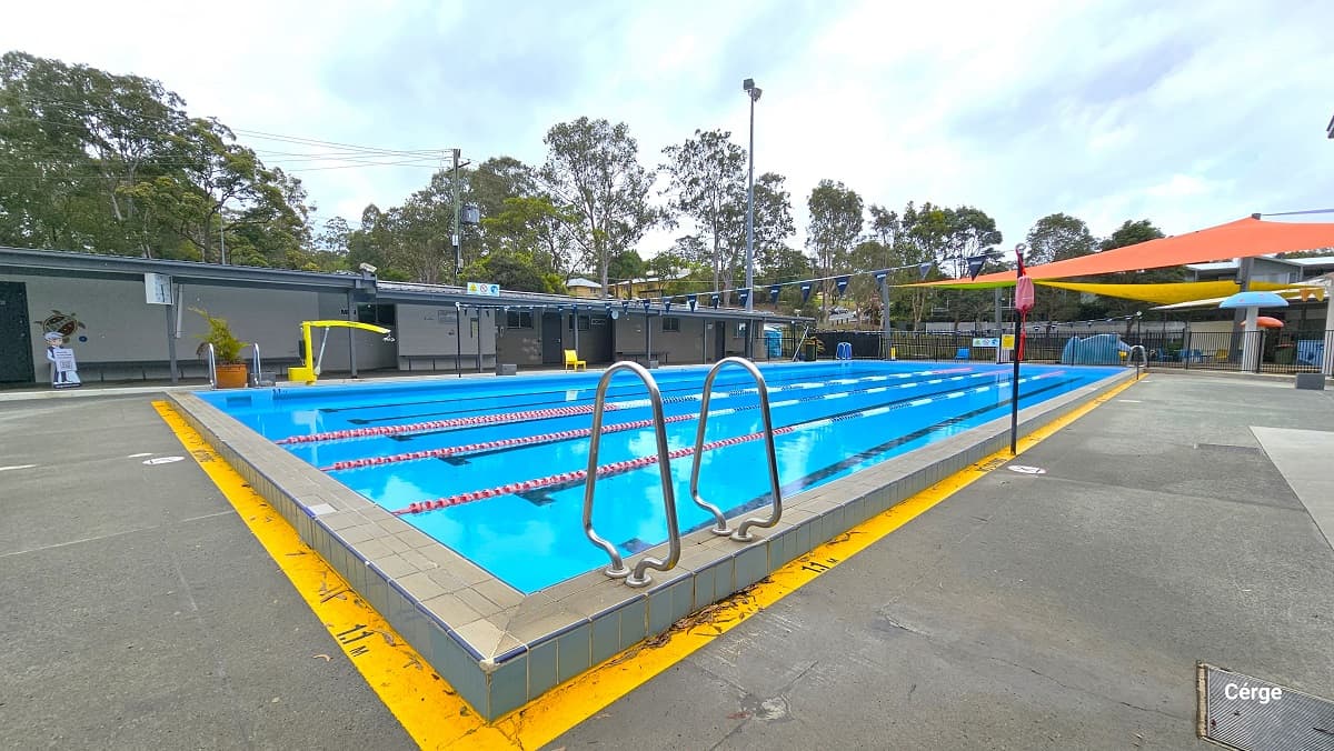 View on the right side of the 25m, 4-lane pool. There is a low-set grey and cream building at the back with long benches placed against the white wall. It has small glass windows for air circulation and illumination. A large, square, white clock can be seen hanging on the roof of the building. The Cérge app boy icon holding a QR code for people with disabilities can be found in front of a grey door near the pool.
