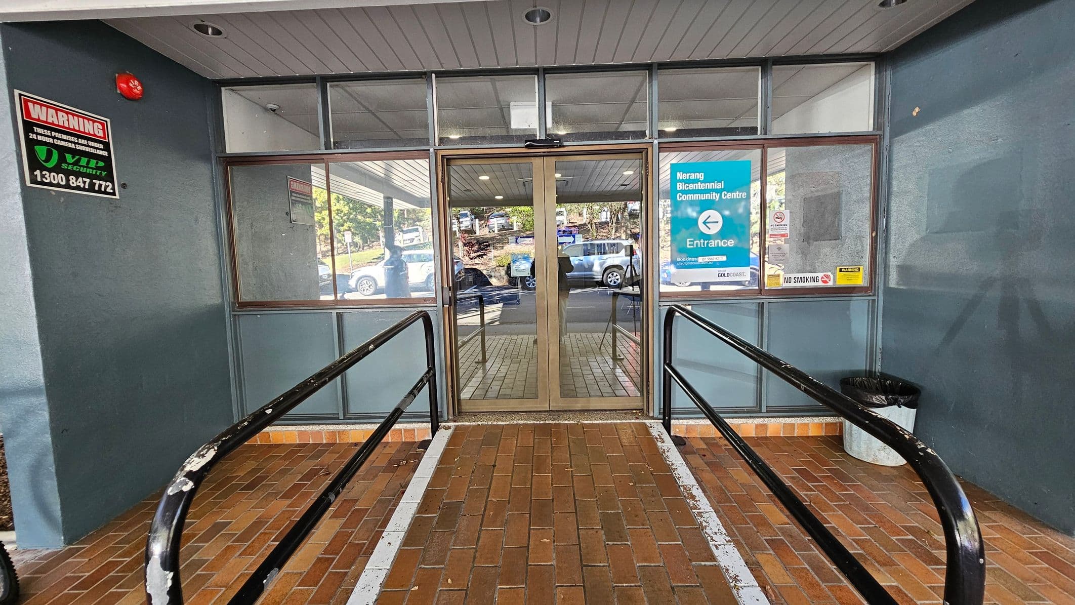 Glass entrance doors with metal frames are in the centre, flanked by large windows. A sign on the right reads "Nerang Bicentennial Community Centre" with an arrow indicating the entrance. A "No Smoking" sign is also visible. On the left, a security warning sign is displayed. The floor is made of brown bricks, and there are black handrails on either side of a ramp leading to the doors. A grey ceiling with recessed lights is above, and a black bin is on the right.