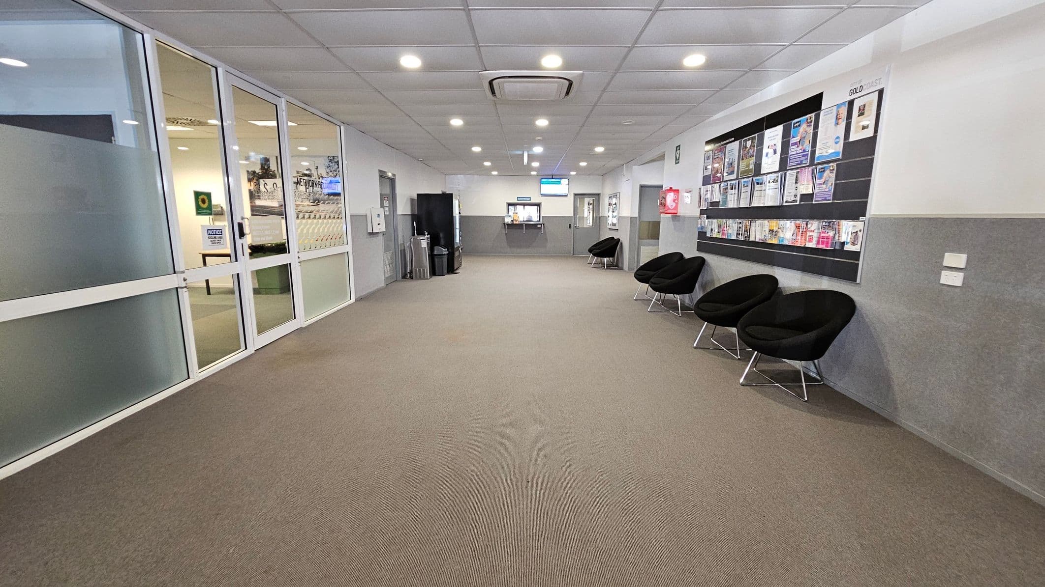 A hallway with a brown carpet and a white tiled ceiling featuring recessed lights. On the left, there are glass doors with a view into an adjacent room. On the right, a wall-mounted bulletin board displays various pamphlets and notices above a row of black chairs. In the background, a counter with a digital screen is visible, flanked by more chairs and a vending machine.
