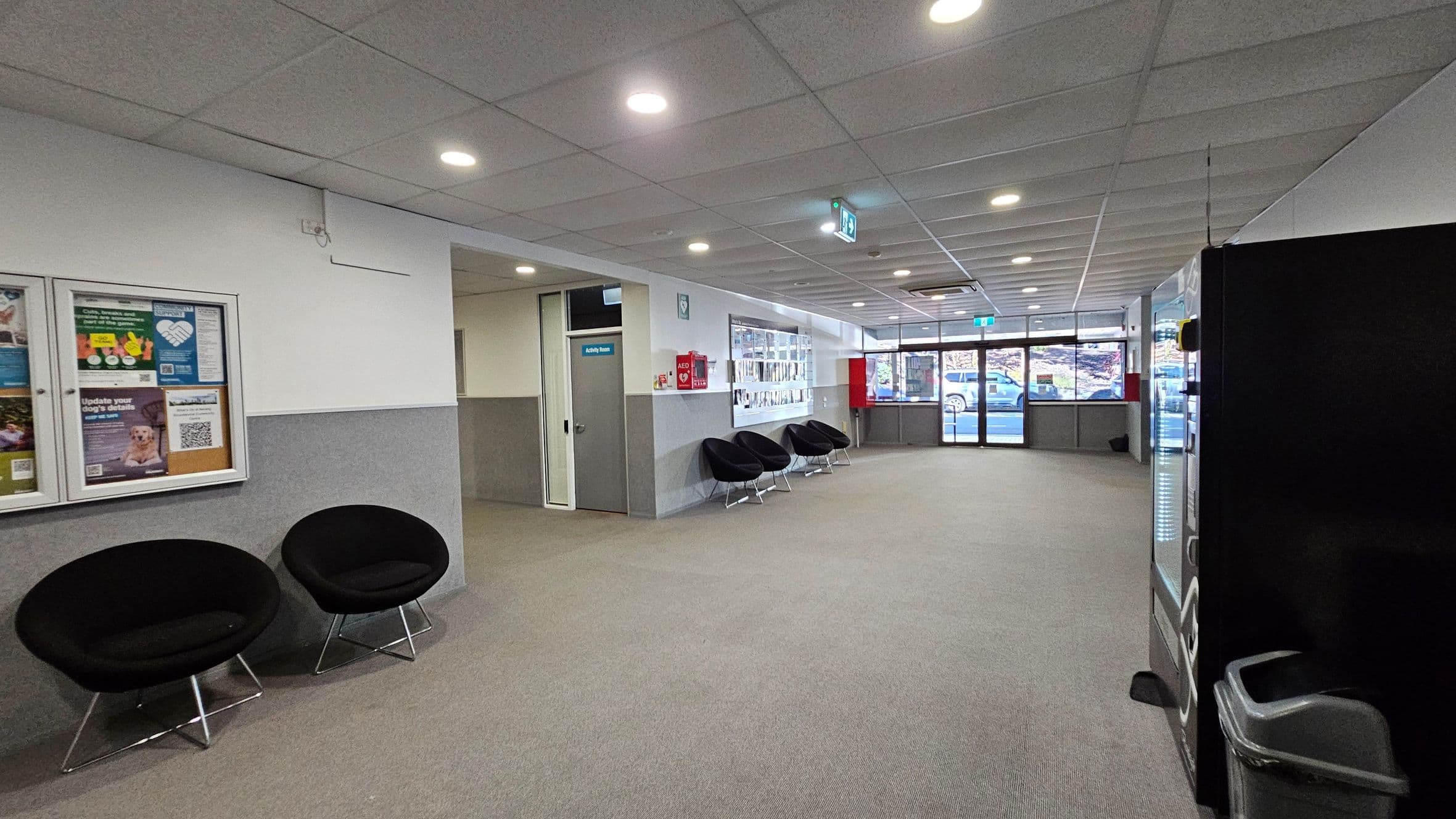 A spacious waiting area with grey carpet and a tiled ceiling with recessed lighting. On the left, there are two black circular chairs and a bulletin board with various notices. In the middle, more black chairs are lined up against the wall. A door labelled "Activity Room" is visible. On the right, a vending machine and a waste bin are present. The background features large windows and glass doors, showing a view of parked cars outside.
